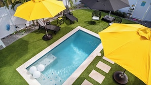 Une petite piscine bordée de carreaux, entourée de gazon synthétique et ombragée par deux parasols jaunes.