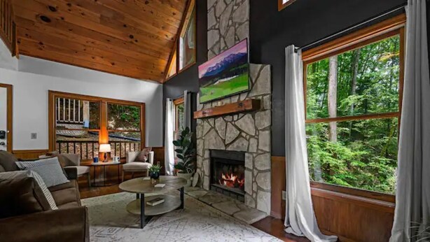 A cozy living room with a stone fireplace, wooden ceiling, and large windows overlooking a forest. - Romantic Smoky MTN Cabin • Hot Tub • Creekside • Near Dollywood • 1mi 2Pkwy (Sevierville)