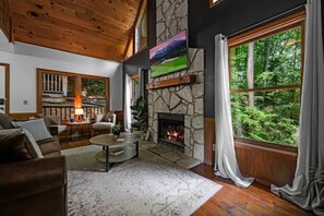 A cozy living room with a stone fireplace, wooden ceiling, and large windows overlooking a forest. - Creek Cabin • Hot Tub • Fireplace • Near Dollywood (Sevierville)
