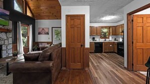 A spacious living area with a wooden ceiling, a stone fireplace, a brown leather sofa, and a kitchen with wooden cabinets and a black dishwasher. - Romantic Smoky MTN Cabin • Hot Tub • Creekside • Near Dollywood • 1mi 2Pkwy (Sevierville)