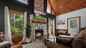 A cozy living room with a stone fireplace, wooden ceiling, and large windows overlooking a forest. - Romantic Smoky MTN Cabin • Hot Tub • Creekside • Near Dollywood • 1mi 2Pkwy (Sevierville)