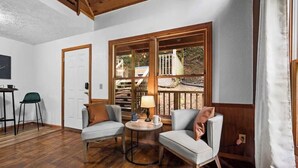 A cozy room with two armchairs, a small table, and a window with a view of a deck and outdoor area. - Romantic Smoky MTN Cabin • Hot Tub • Creekside • Near Dollywood • 1mi 2Pkwy (Sevierville)