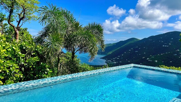 Uma piscina infinita azul límpida, com borda em azulejo, rodeada por vegetação exuberante e com vista para a costa montanhosa.