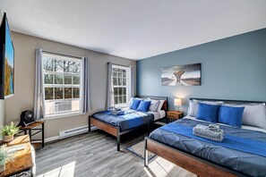 Two single beds with blue bedding and white pillows, a reading nook with a fan and potted plant, and a framed artwork adorn this serene bedroom.