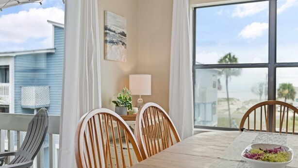 A dining area with a table, chairs, a bowl of salad, a lamp, and a view of the beach through large windows. - Ocean View/Deeded Beach Access/Selfie Wall/Pets Ok! (Gulf Shores)