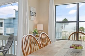 Ein Essbereich mit Tisch, Stühlen, einer Salatschüssel, einer Lampe und Blick auf den Strand durch große Fenster.