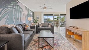 A modern living room with a grey sofa, a coffee table, and a TV mounted on the wall. - Ocean View/Deeded Beach Access/Selfie Wall/Pets Ok! (Gulf Shores)