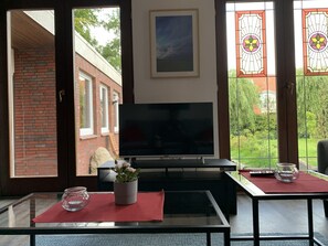 Uma sala de estar com televisão, mesa de centro de vidro e planta em vaso. Há duas cadeiras e uma mesa com toalha vermelha. O quarto tem uma parede de tijolos e uma janela com vista para um jardim.