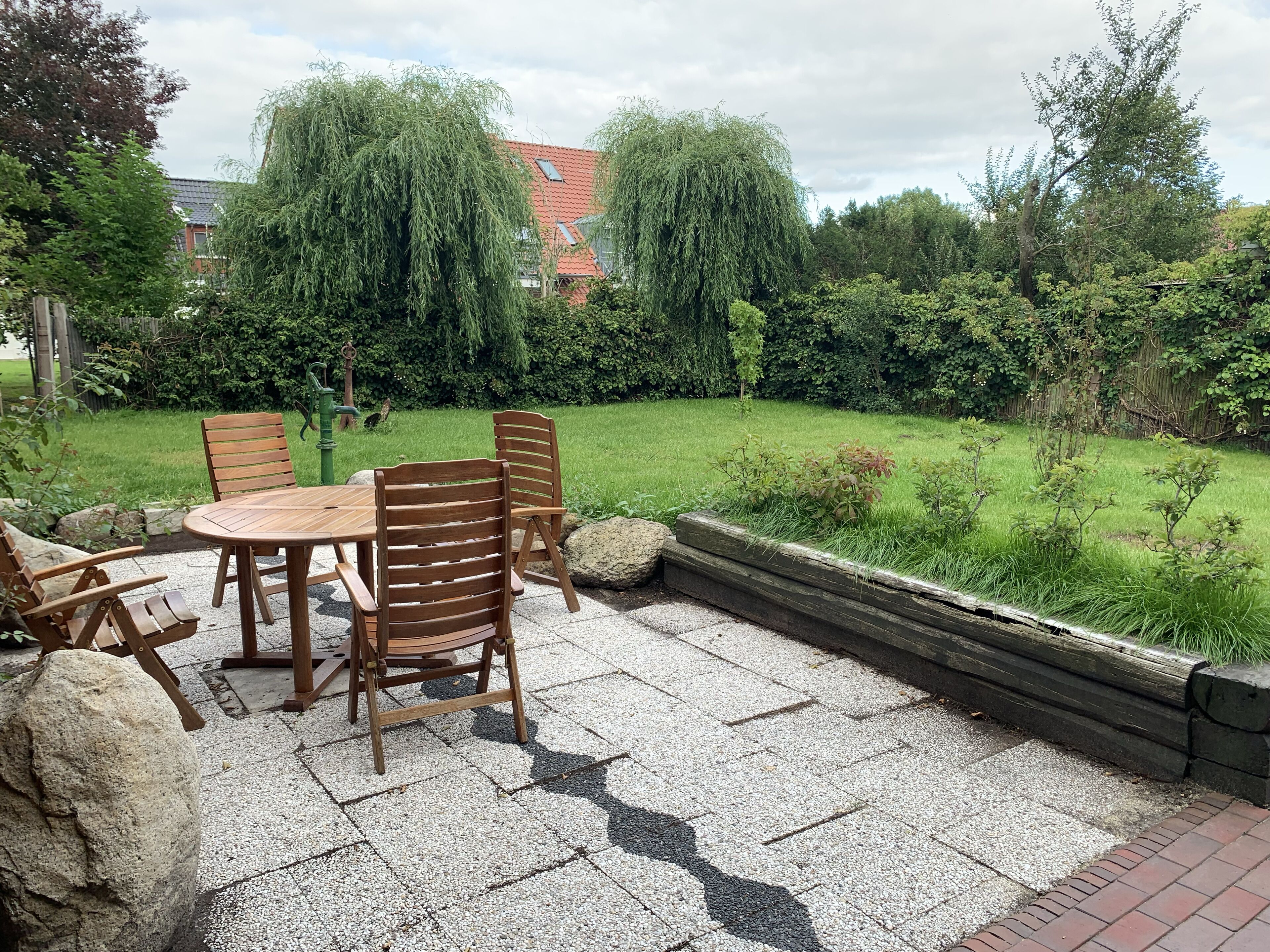 An outdoor patio with a round wooden table and chairs, surrounded by a gravel floor and a low stone wall.