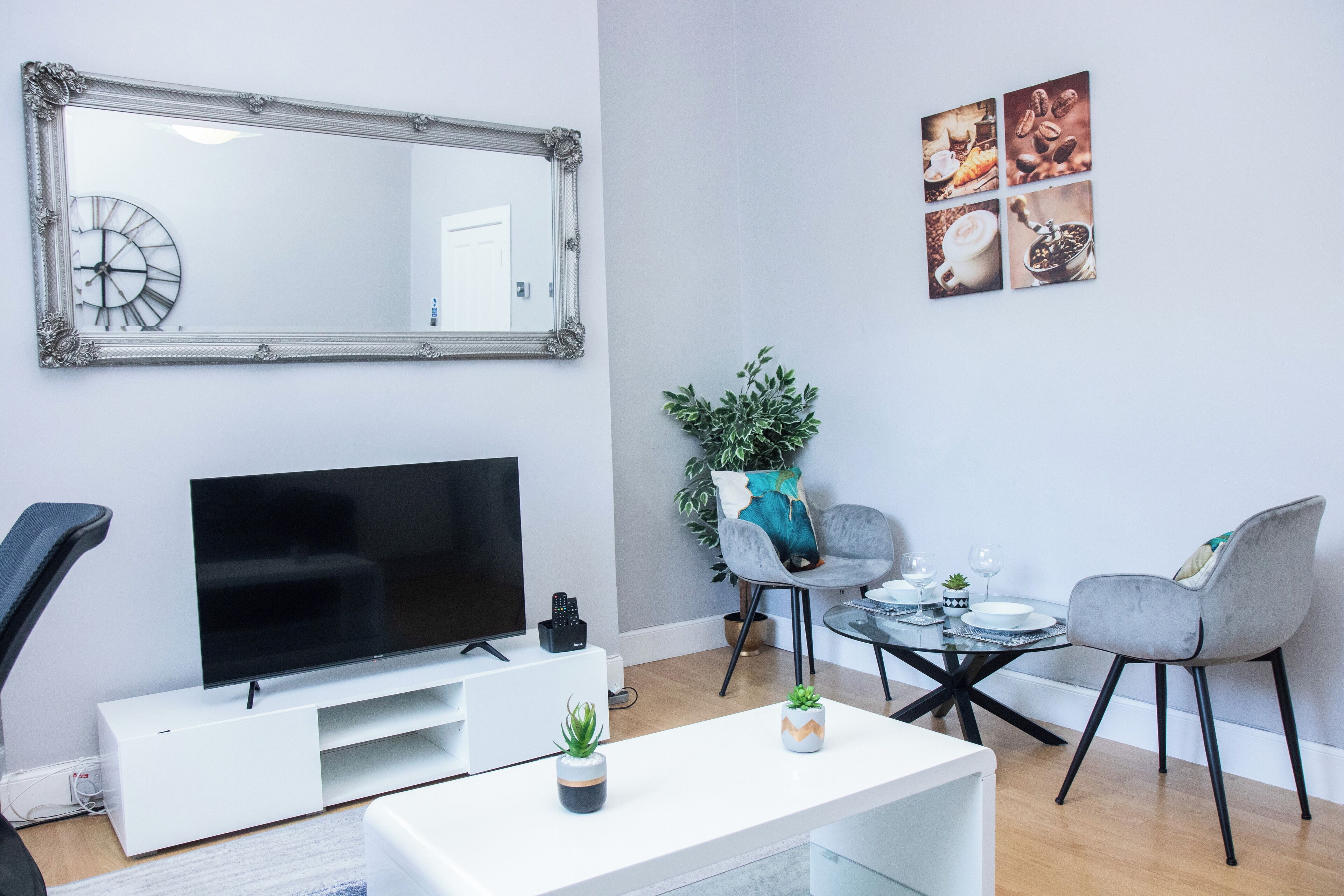 Ein modernes Wohnzimmer mit Flachbildfernseher, weißer Unterhaltungseinheit, zwei grauen Sesseln, einem Couchtisch und einem großen Spiegel mit Uhr.