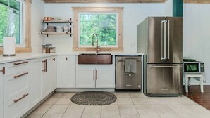 A modern kitchen with white cabinets, a stainless steel refrigerator, and a window with a view of greenery.