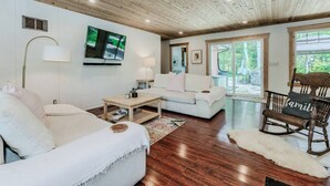 A modern living room with a white sofa, a wooden coffee table, a white armchair, and a rocking chair. There is a television mounted on the wall and a window providing a view of the outdoors.