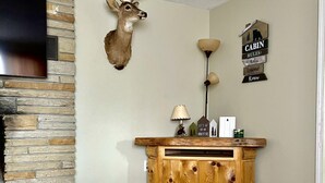 Una sala de estar con chimenea de piedra, una cabeza de venado montada, un mueble de madera, un televisor y un cartel que dice 'CABIN RULES'.