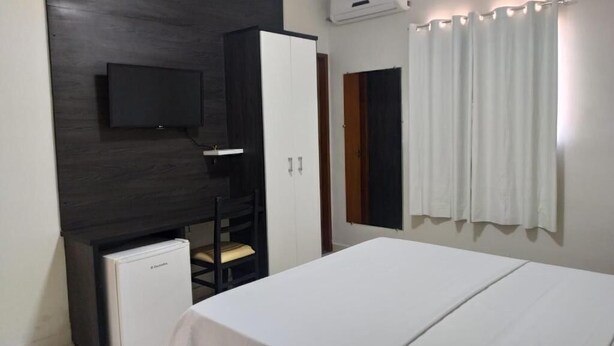 Un dormitorio con cama, televisión montada en la pared, armario, refrigerador pequeño y ventana con cortinas.