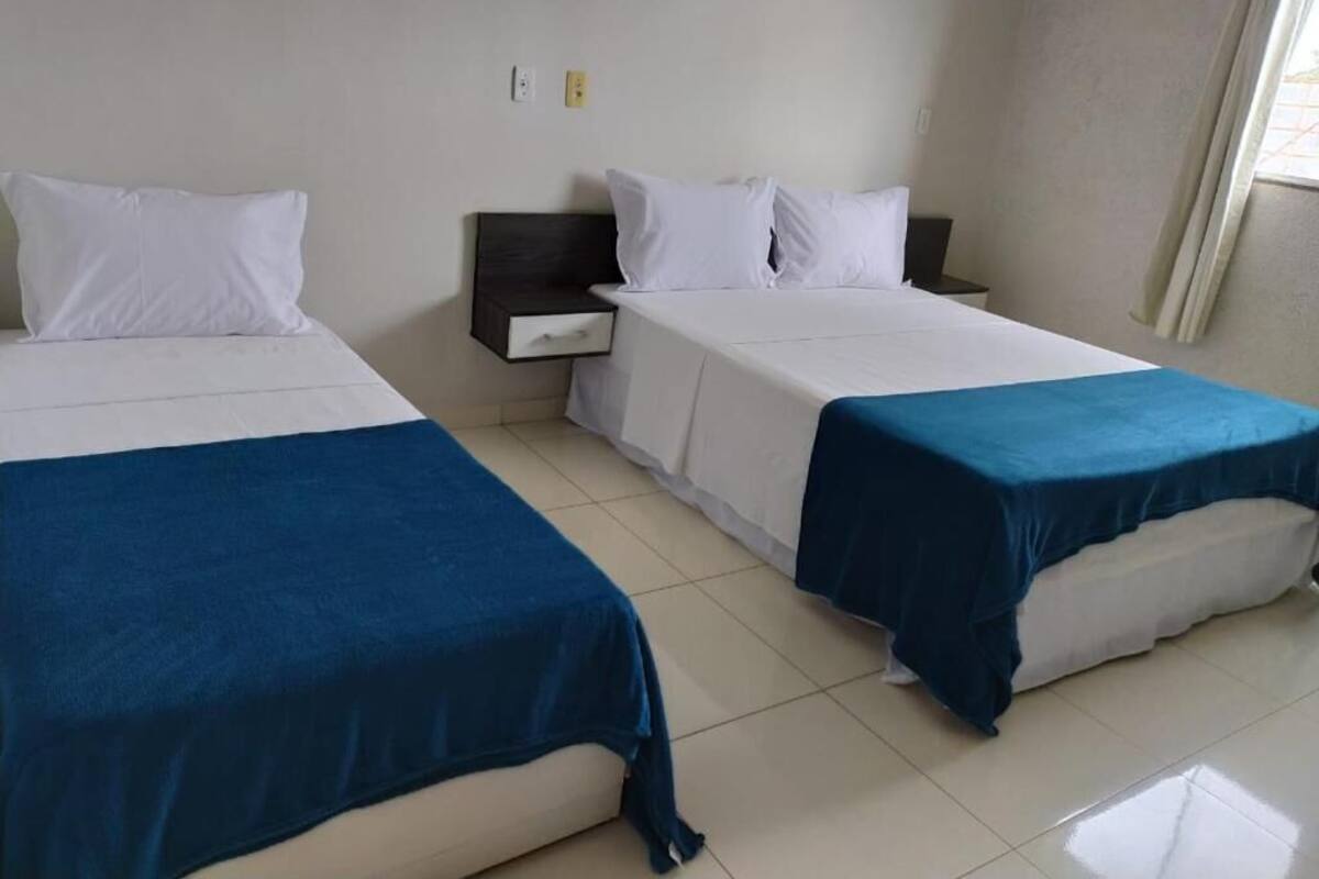 A hotel room with two beds, each with a white mattress and blue bedspread. There is a nightstand between the beds with a dark-colored drawer. The room has white walls and a window with curtains.