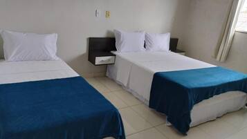 Habitación de hotel con dos camas, cada una con colchón blanco y colcha azul. Hay una mesita de noche entre las camas con un cajón oscuro. Las paredes son blancas y hay una ventana con cortinas.