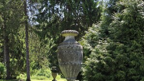 Un jardín con una urna de piedra grande, césped verde y algunas flores púrpuras.