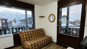 Un cuarto acogedor con un sofá de cuadros, una pequeña mesa de madera y ventanas amplias que ofrecen vistas a edificios cubiertos de nieve y montañas.