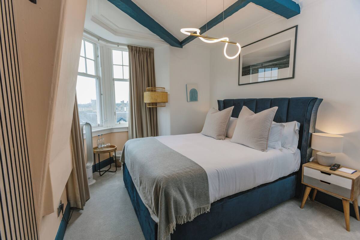 A bedroom with a large bed, a bedside table with a lamp, a window with curtains, and a city view.