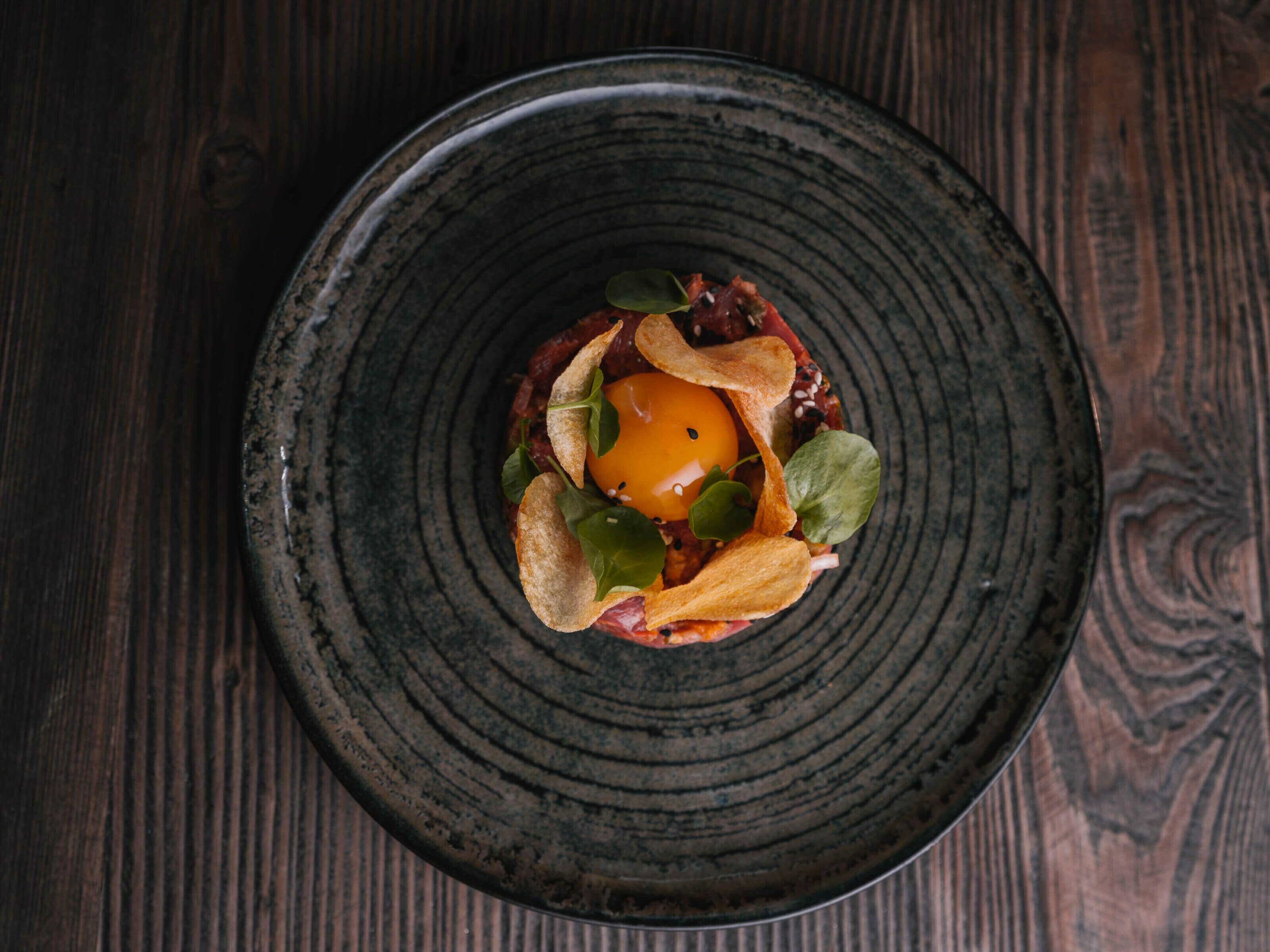 A dark ceramic plate with a central egg yolk surrounded by greens and thinly sliced meat, placed on a wooden table.