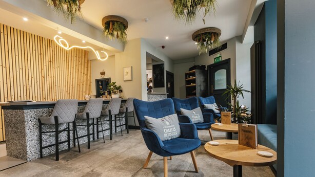 A modern bar area with blue upholstered chairs, a wooden bar counter, and hanging plants. - Haar with Rooms (St. Andrews)