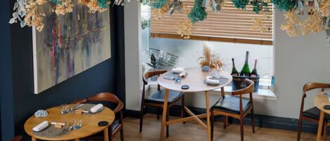 Un espace repas avec des tables et des chaises en bois, une grande peinture abstraite au mur et une fenêtre avec des stores.