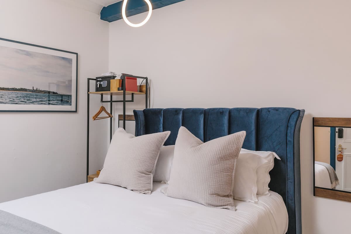 A neatly made bed with white bedding and pillows, a blue upholstered headboard, a framed picture of a coastal scene, and a bedside shelf with books and a phone.