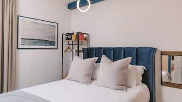 A neatly made bed with white bedding and pillows, a blue upholstered headboard, a framed picture of a coastal scene, and a bedside shelf with books and a phone.