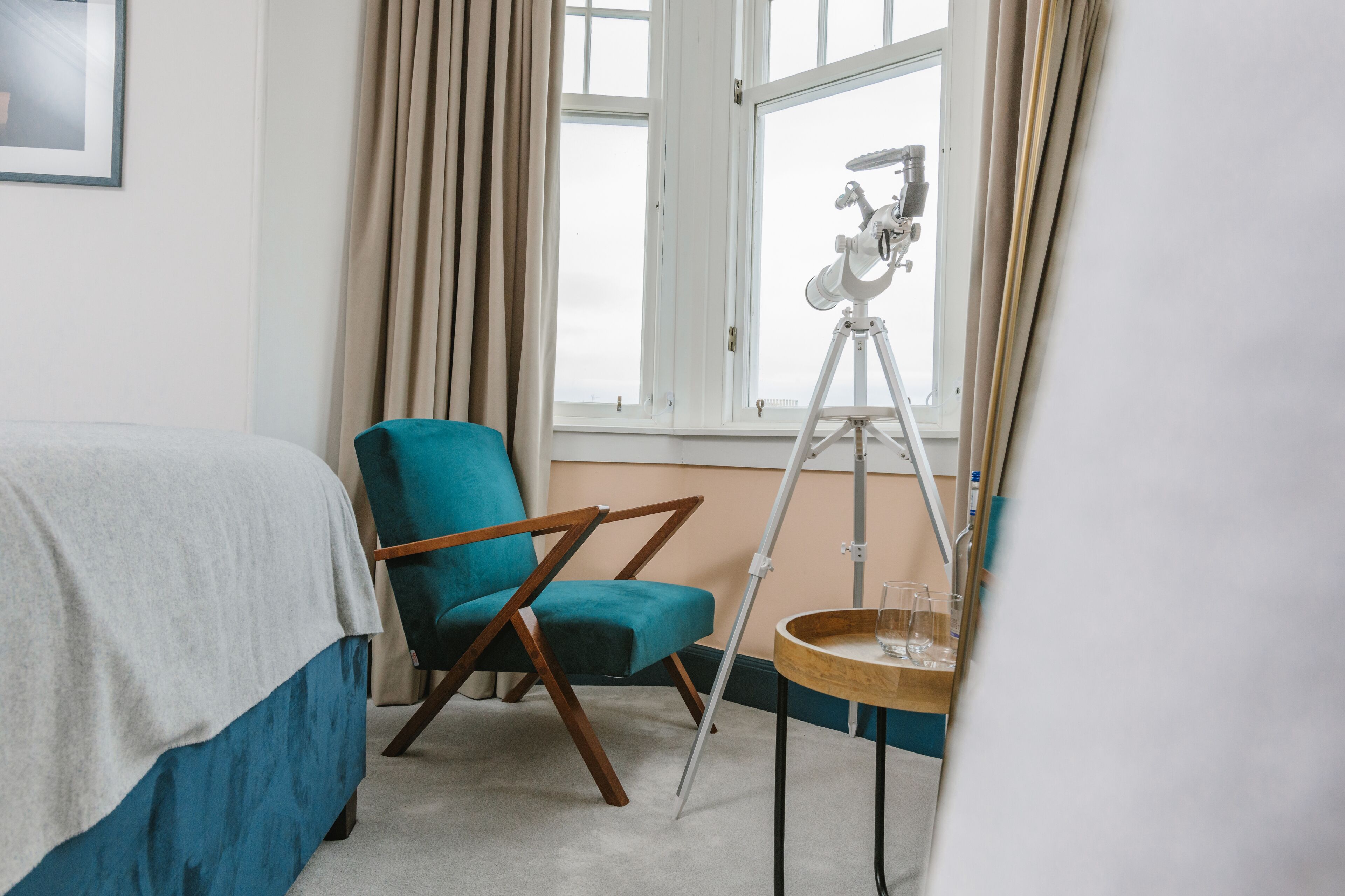 A bedroom with a bed, a teal armchair, a telescope, and a small table with glasses.
