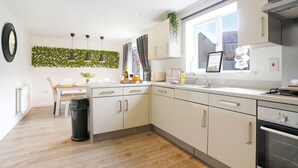 A modern kitchen with white cabinets, a black trash bin, a dining table with chairs, and a window with curtains. - Detached House • Free Parking • FastWifi • SmartTV (Northampton)