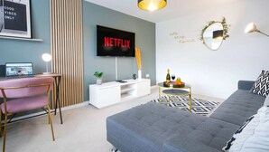 A modern living room with a grey sofa, a TV displaying 'NETFLIX', a desk with a laptop, and a small round table with drinks. - Detached House • Free Parking • FastWifi • SmartTV (Northampton)