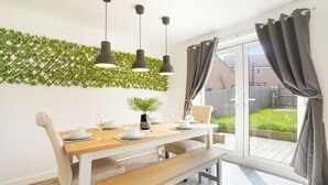 A dining area with a wooden table, white chairs, and a bench. There are three pendant lights above the table. A green plant is on the table, and a window with curtains is visible. - Detached House • Free Parking • FastWifi • SmartTV (Northampton)