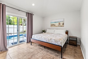 Un dormitorio con una cama, una mesita de noche pequeña, un cuadro en la pared y una puerta corrediza de vidrio que da a una zona de piscina.