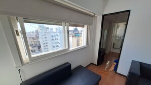 Une pièce dotée d’une grande fenêtre, d’un canapé noir et d’une salle de bain visible à travers une porte ouverte.