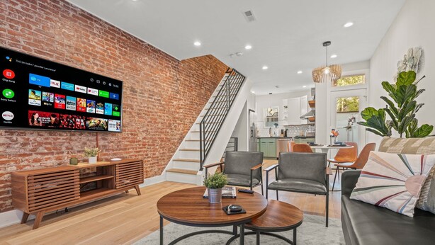 A modern living room with a brick wall, a wooden entertainment unit, a round coffee table, and a black leather sofa. - Clean and Comfy | King Bed | Steps to Fishtown Fun (Philadelphia)
