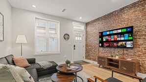 A modern living room with a brick accent wall, a flat-screen TV displaying streaming services, a black leather sofa, a round coffee table, and a wooden entertainment unit. - Clean and Comfy | King Bed | Steps to Fishtown Fun (Philadelphia)