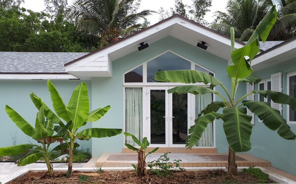 Una casa con fachada azul claro, puerta principal blanca y ventanas amplias. Hay plantas de plátano en el frente.