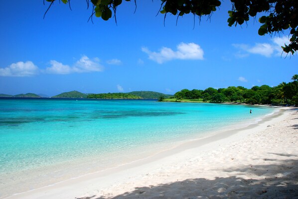 A beach with clear turquoise water, white sand, and green hills in the background.