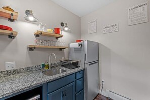 A kitchen with a granite countertop, a sink, a refrigerator, and wooden shelves with glassware.