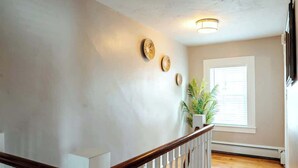A staircase with a wooden handrail and white balusters, a wall with three circular wall art pieces, a potted plant, and a window with blinds. - Charming & Historic 2BR Oasis in Downtown Luxury (Manchester)