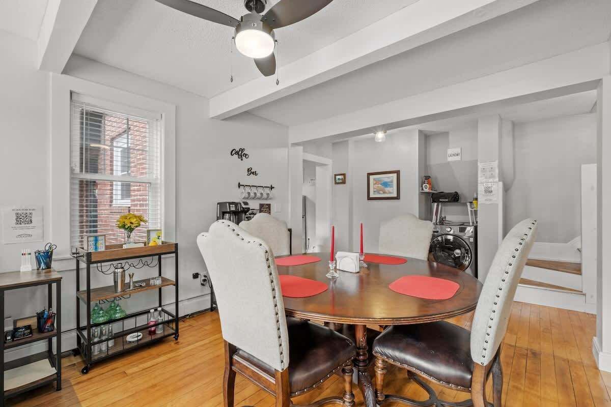 A dining area with a round table, four chairs, a coffee bar, and a window with blinds.
