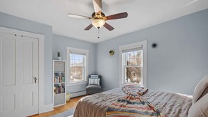 A bedroom with a bed, ceiling fan, windows, and a bookshelf. - Charming & Historic 2BR Oasis in Downtown Luxury (Manchester)