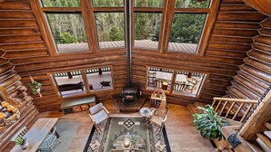 A log cabin with a central fireplace, wooden furniture, and large windows offering a forest view.