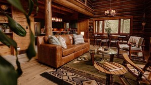 A cozy log cabin interior with a leather sofa, dining table, and chairs. A kitchen area is visible in the background.
