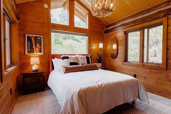 Un dormitorio de cabaña de madera con una cama, mesitas de noche y un ventanal grande.