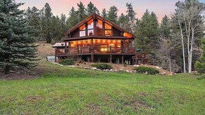 A two-story log cabin with a deck and large windows, surrounded by trees and a grassy area.