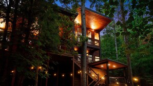A multi-level wooden cabin with a staircase and string lights, surrounded by trees. - Cozy Nights Cabin Luxury and Location (Broken Bow)