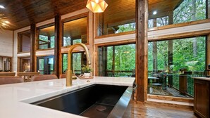 A modern kitchen with a large island, wooden beams, and large windows overlooking a forested area. - Cozy Nights Cabin Luxury and Location (Broken Bow)
