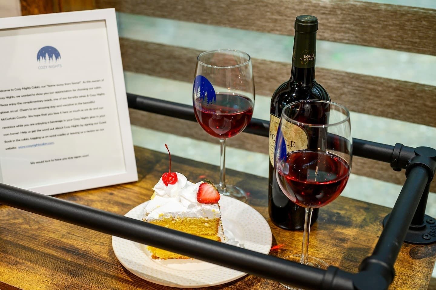 Una mesa de madera con una porción de pastel cubierta de crema batida y una cereza, dos copas de vino tinto y una botella de vino.