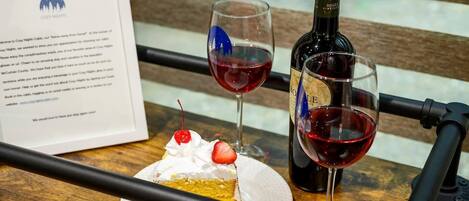 Una mesa de madera con una porción de pastel cubierta de crema batida y una cereza, dos copas de vino tinto y una botella de vino.
